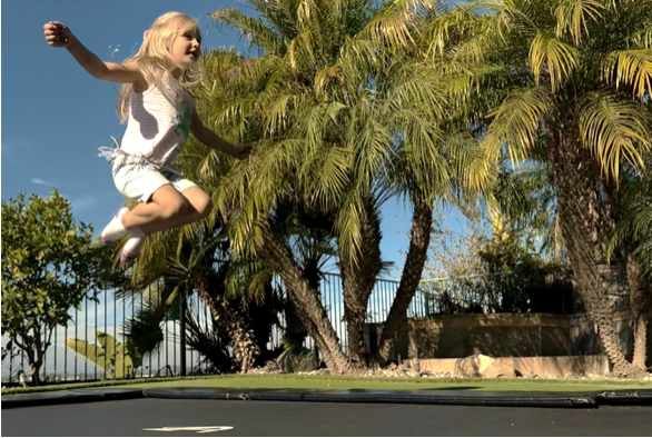 A young girl joyfully jumps on a trampoline in a sunny backyard, surrounded by green grass and colorful flowers.